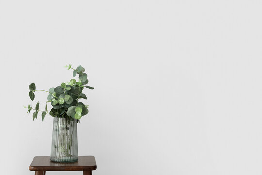 Vase with eucalyptus on wooden stool near light wall