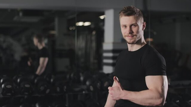 The athlete is standing in the gym near a dumbbell rack. He is turning his head and looking at the camera. Training in the gym. 4K