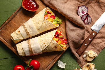 Board of tasty Mexican burritos with vegetables on green wooden background