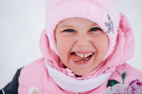 Closeup photo of kid smiling and sticking tongue out of mouth in a funny way wearing warm winter clothes in forest. Astonishing background full of white color and snow. 