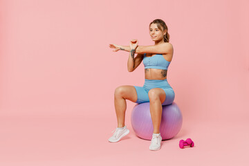 Fototapeta na wymiar Full body young athletic fitness trainer instructor woman wear blue tracksuit spend time in home gym sit on fitball stretch hands isolated on pastel plain light pink background Workout sport concept.