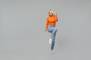 Full body elderly overjoyed excited happy blonde woman 50s in orange turtleneck do winner gesture raise up leg clench fist isolated on plain grey background studio portrait. People lifestyle concept. - Powered by Adobe