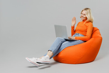 Full body elderly smiling blonde woman 50s in orange turtleneck sit in bag chair hold use work on laptop pc computer waving hand talk by video call isolated on plain grey background studio portrait