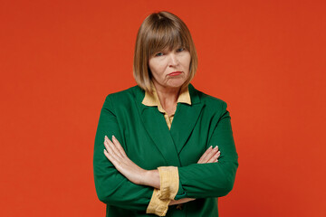 Elderly sad strict angry dissatisfied displeased caucasian woman 50s wearing green classic suit hold hands crossed folded isolated on plain orange color background. People business lifestyle concept.