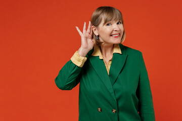 Elderly fun curious nosy woman 50s wearing green classic suit try to hear you overhear listening intently isolated on plain orange color background studio portrait. People business lifestyle concept.