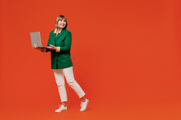 Full body elderly happy smiling woman 50s wearing green classic suit hold use work on laptop pc computer look aside isolated on plain orange color background studio. People business lifestyle concept.