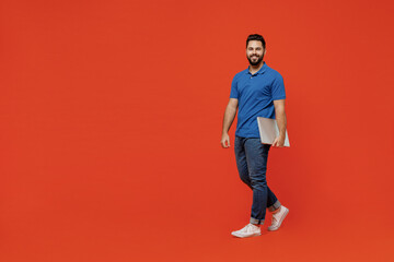 Fototapeta na wymiar Full body young smiling confident happy caucasian man 20s wear basic blue t-shirt looking camera hold closed laptop pc computer walk go isolated on plain orange background People lifestyle concept