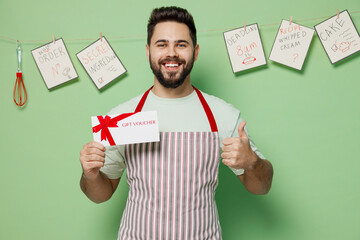 Young chef confectioner baker man in striped apron hold store gift certificate coupon voucher card show thumb up isolated on plain pastel light green background studio portrait. Cooking food concept.