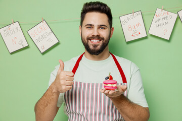 Young male chef confectioner baker man 20s in striped apron hold pink little cake muffin macaroon show thumb up isolated on plain pastel light green background studio portrait. Cooking food concept.