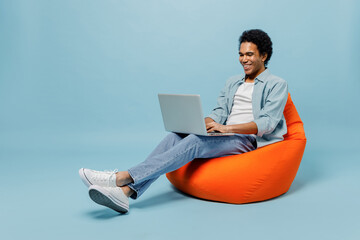 Full size body length young black curly man 20s wears white shirt sit in bag chair hold use work on laptop pc computer typing browsing isolated on plain pastel light blue background studio portrait.