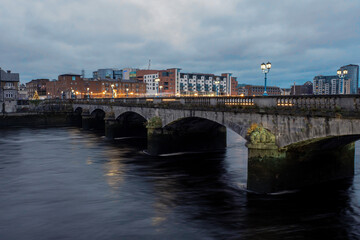 Naklejka premium Riverside area at night. Shannon river, Limerick city, Ireland. Blue and orange colors. Dusk scene. Cloudy sky. Christmas and New Year illumination.