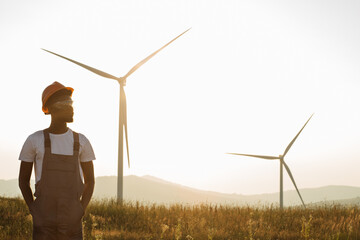 African american man standing on field with huge windmills during summer sunset. Professional technician wearing grey overalls, orange helmet and safety glasses.