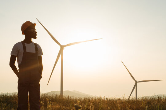 African american man standing on field with huge windmills during summer sunset. Professional technician wearing grey overalls, orange helmet and safety glasses.