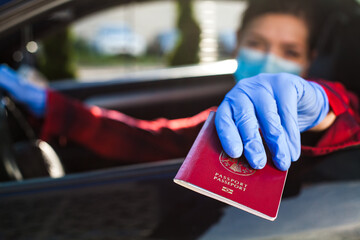 Young female driver wearing protective face mask and gloves giving red passport through car window