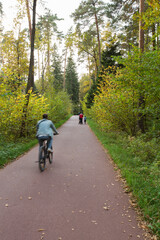 Obraz premium man riding bicycle, woman with son and baby trolley walking on asphalt walkway trail path road in the woods with green grass, trees around. Meshersky forest park. Moscow, Russia