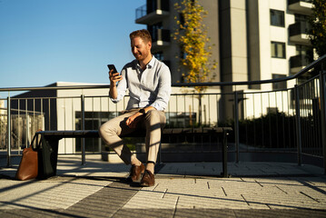 Young stylish businessman sitting on the beanch outdoors. Portrait of handsome man using the phone.