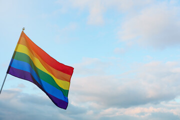 Bright LGBT flag against blue sky with clouds. Space for text