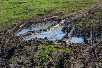 Mud puddle road spring. A rural road in a green field. Spring snow melting, the first greenery. The concept of warm spring days. Off-road driving. Puddles with a reflection of the sky. Car tracks