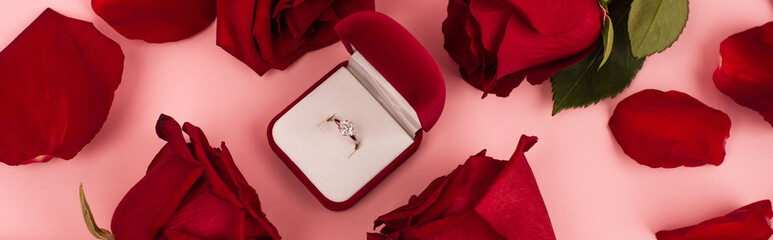 flat lay of red rose petals around jewelry box with diamond ring on pink, banner.
