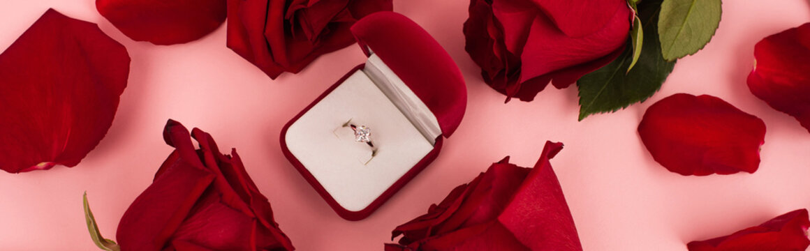 flat lay of red rose petals around jewelry box with diamond ring on pink, banner.