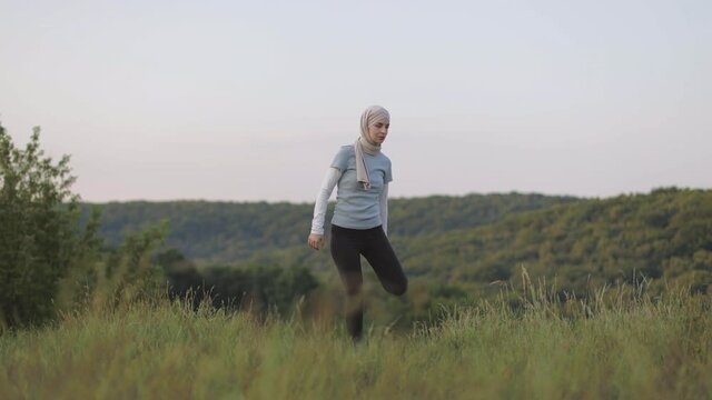 Active woman in sport clothes and hijab stretching legs at summer park. Young arabian man standing on background and doing physical exercises Sport activity outdoors.
