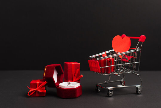 Jewelry Box With Diamond Ring Near Presents And Small Shopping Cart On Black.