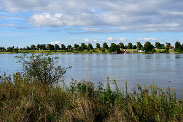 A view of a vast yet shallow river connecting two sides of a peninsual with a road ending next to a post seen on a cloudy summer day on a Polish countryside during a hike