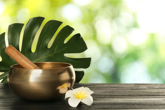 Golden singing bowl, mallet, green leaf and flower on wooden table outdoors, space for text