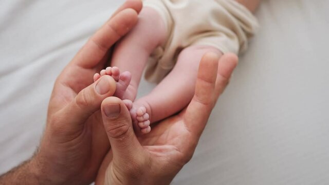 Father holding tiny feet of newborn baby in his hands and petting them. Parent of adorable infant cares about his child at home. Beautiful moments of fatherhood