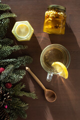on a wooden table there is a jar of honey with nuts, an open jar of cream honey, tea with lemon, a wooden spoon and branches of a Christmas tree.
