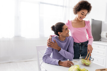 African american woman hugging boyfriend and cooking salad in kitchen.