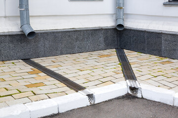 Obraz premium facade of a building wall with a downspout pipe and a concrete dish canal with iron grid on a stone slab sidewalk to drain rainwater near asphalt road city street, nobody.