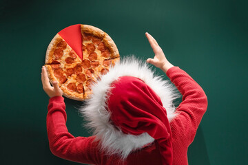 Top view of Pepperoni pizza and back of boy in red santa claus hat with fluffy fur  and on green background. The concept of a holiday, a Christmas Time to eat