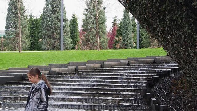 A teenage girl walks near the stairs, along which water flows down from above against the background of a green park. A girl in a gray jacket walks against the background of water pouring from above