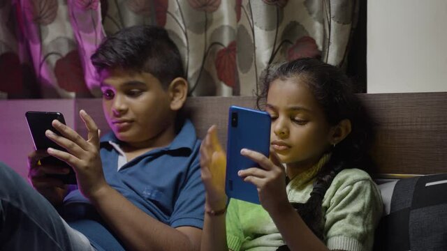 Two Young Teenage kids busy using mobile phone at night on bedroom while sleeping - concept of children smartphone and technology addiction.