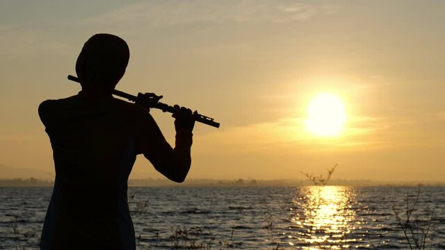 silhouette of man playing flute with water and sunrise background. Slow motion. 