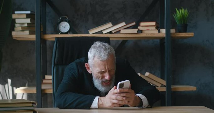 Angry mature businessman reacts aggressively to bad message using smartphone while sitting in office. Bearded male entrepreneur bangs his fist on the table, reaction to failure