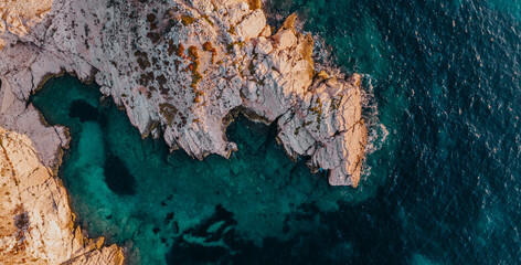 Panoramic top down view of a rocky coast and deep blue crystalline waters during sunset. 