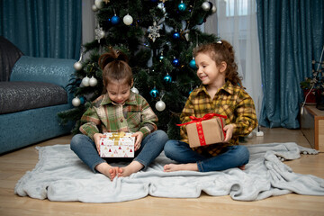 Obraz na płótnie Canvas Two cute beautiful girls sit near the Christmas tree with gifts. New Year. Christmas.