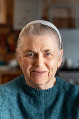 The face of a happy elderly woman of the 80s. He smiles and looks at the camera. Portrait of an old lady with gray hair in a knitted green jacket. There is a blurry image of the kitchen.