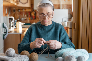 An elderly lady of 80 years old is engaged in an interesting hobby, knitting a brown jacket with knitting needles. Hobbies that you can do at home. A blurry image of the kitchen.