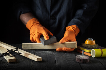 Woodworker cleans a wooden plank with an abrasive tool. Hands of the builder close-up during work. Renovation or construction idea.