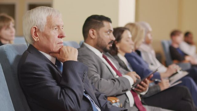 PAN shot of Caucasian senior man and other political leaders of different countries listening to speaker at conference, sitting in row in big auditorium