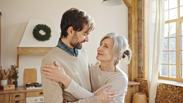 Tracking shot portrait of loving adult son embracing his senior grey-haired mother, smiling together to camera at home