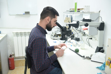 A young man looks through a microscope examining repairs dentures or a jaw in the workshop of a dental technician