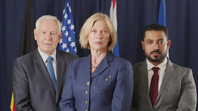Tilt up medium slowmo portrait of two male and one female diverse political leaders looking at camera standing on dark blue background with un flags