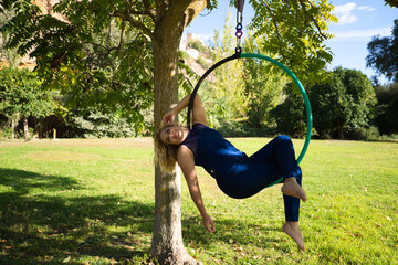 Blonde woman and young gymnast acrobat athlete performing aerial exercise on air ring outdoors in park. Lithe woman in blue costume performs poses of circus performers dancing with hips