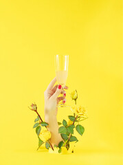 Woman's hand holds a glass of champagne and comes out along with yellow rosses from the background....