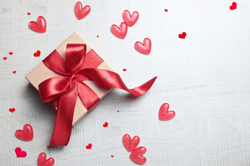 Top view photo of valentine's day decorations white gift box with red ribbon bow and small hearts on light background.