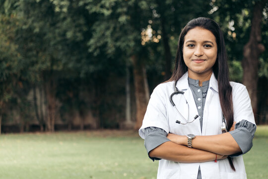 Portrait Of Confident Indian Doctor Wear White Coat, Standing At Outdoors With Arms Crossed On Chest. Professional Front Line Covid Warriors During Covid-19, Medical Service Concept. With Copy Space